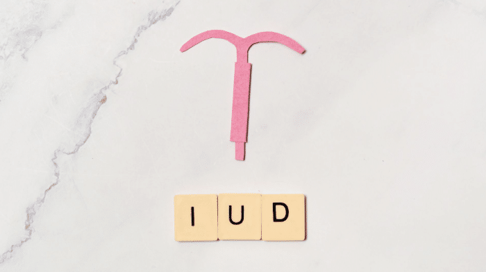 A pink intrauterine device (IUD) symbol made out of paper is placed above three beige tiles with black letters spelling "IUD" on a marble background.