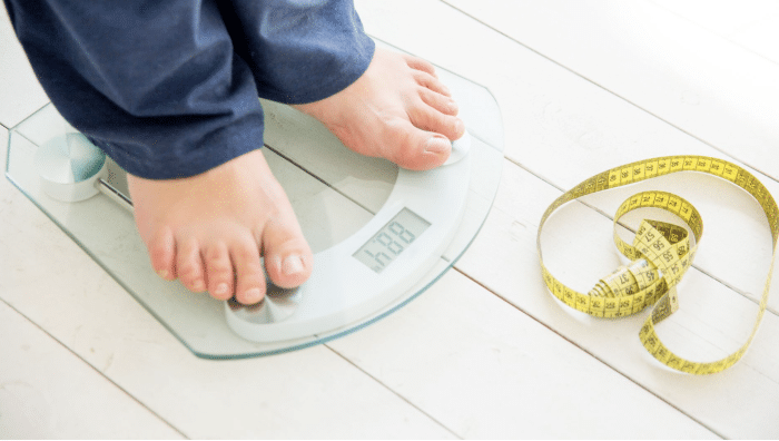 A person standing on a digital scale displaying a weight of 68.8. Next to the scale, there is a yellow measuring tape lying on a white wooden floor. The person is wearing dark blue pants.