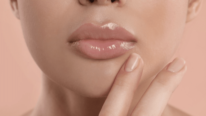 Close-up of a person's glossy, pink lips and part of their chin. The person is gently touching their chin with a polished fingernail. The background is a soft peach color.