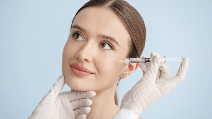 A woman with light skin and brown hair tied back is receiving a cosmetic injection in her cheek. She looks upward with a gentle smile. Gloved hands are holding her chin and administering the syringe. The background is a light blue.