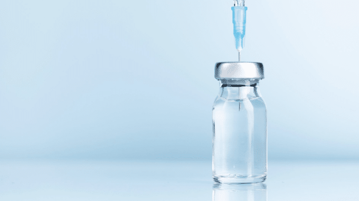 A close-up view of a clear glass vial with a silver lid, filled with a liquid, positioned on a reflective surface. A syringe with a blue needle is inserted into the top of the vial against a light blue background.