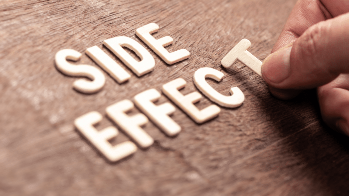 A close-up of a hand arranging wooden letters on a wooden surface to spell out the words "SIDE EFFECT." The letters appear to be in a scattered arrangement with the hand placing the last "T" in "EFFECT.