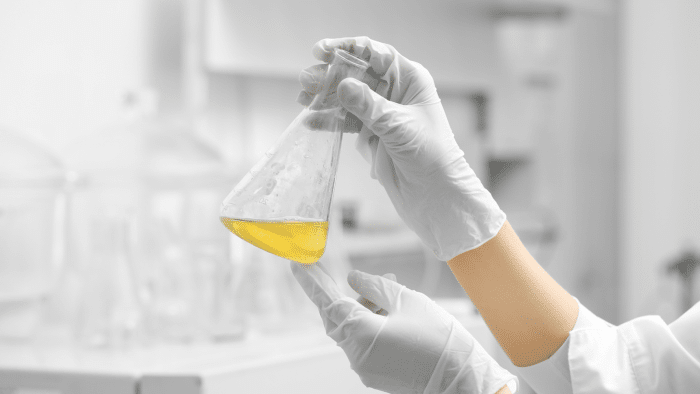 A person wearing white gloves and a white lab coat holds up a clear Erlenmeyer flask containing a yellow liquid in a bright, sterile laboratory setting. Various lab equipment can be seen blurred in the background.