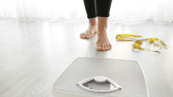 A person standing barefoot on a wooden floor, approaching a white analog bathroom scale. A yellow measuring tape is coiled on the floor nearby. The room is lit naturally with sheer curtains in the background.