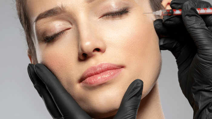 A person with closed eyes receives an injection in the cheek from a gloved hand holding a syringe. The person appears calm, with the other gloved hand gently supporting their face.