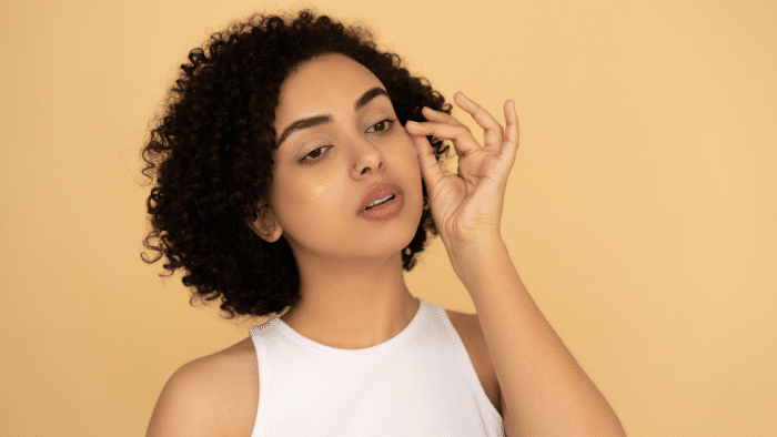 A person with curly hair and wearing a sleeveless white top applies a facial patch under their eye with a gentle touch. The background is a solid beige color.