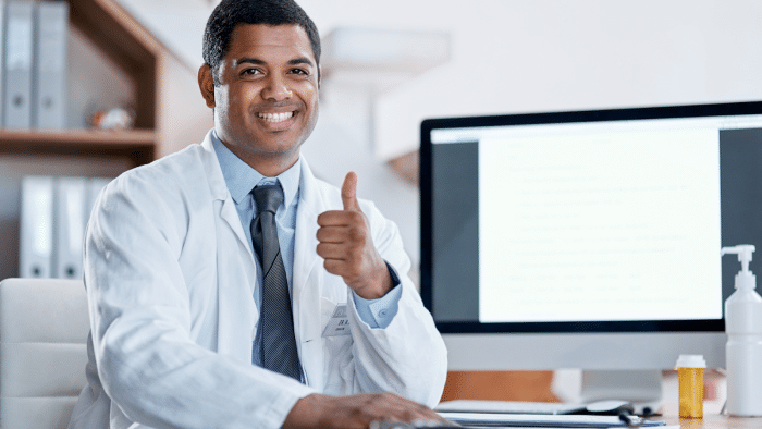 A doctor in a white lab coat and tie sits at a desk, smiling and giving a thumbs-up. Behind him is a large computer monitor. The background includes shelves with binders and office supplies. A bottle of hand sanitizer is visible on the desk.