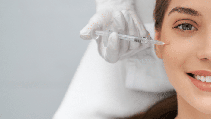A close-up of a woman receiving an injection in the area below her eye. She is smiling while a gloved medical professional's hand administers the injection. Only the right side of the woman's face is visible.