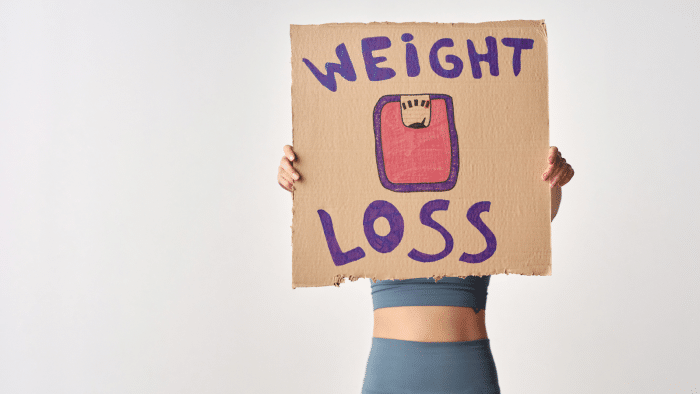 A person wearing blue athletic wear holds up a cardboard sign reading "WEIGHT LOSS" with a drawing of a scale, obscuring their face. The background is white and plain.