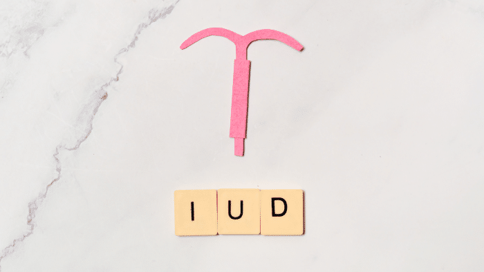 A pink paper cutout of an intrauterine device (IUD) is placed above three yellow tiles with black letters spelling "IUD" on a white marble background.