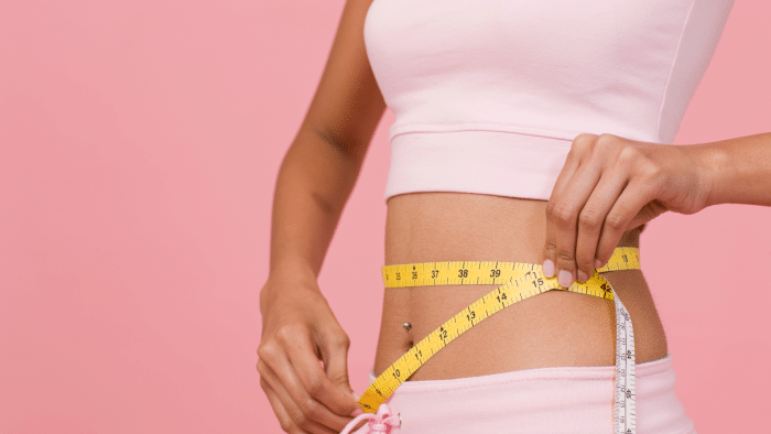 A person wearing a light pink crop top and shorts measures their waist with a yellow measuring tape against a pink background. The focus is on the midsection, with one hand holding the tape in place and the other adjusting it.