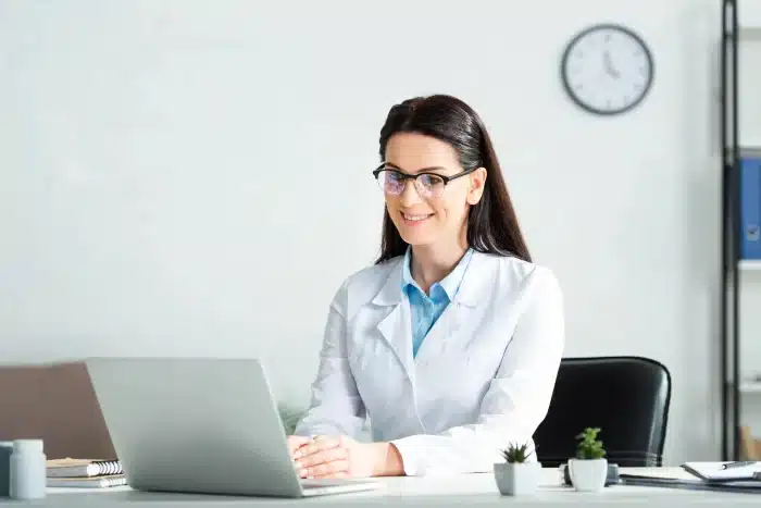 A woman in a white lab coat and glasses is sitting at a desk, smiling at a laptop. There is a clock on the wall behind her and some office supplies and small plants on the desk. She appears to be in a professional, well-lit office setting.