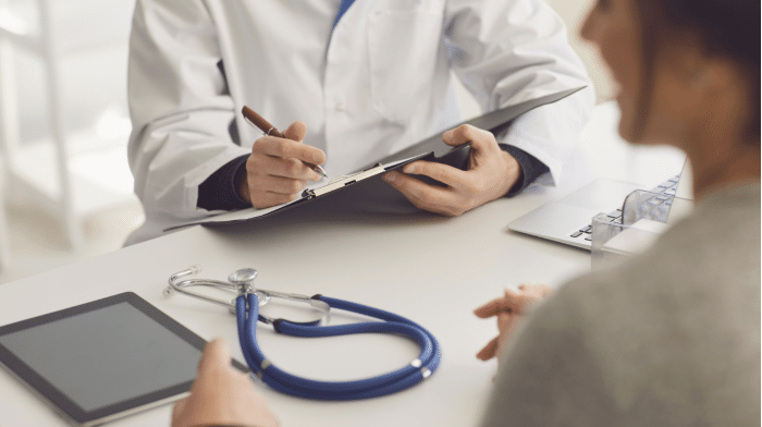 A doctor in a white coat is holding a clipboard and writing notes while talking to a patient. A stethoscope, a tablet, and a laptop are on the desk between them. The image focuses on their hands and objects, not their faces.
