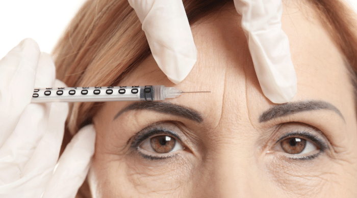 Close-up of a person with gloved hands administering a syringe injection into the forehead. The person's face shows visible wrinkles, indicating a cosmetic procedure, likely Botox or a similar treatment, aimed at reducing the appearance of fine lines or wrinkles.