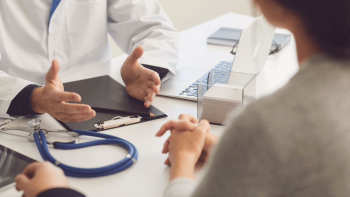 A doctor in a white coat sits at a desk, talking to a patient. The desk has a stethoscope, a clipboard, and a laptop. The patient's hands are clasped, and tissue box is visible in the background.
