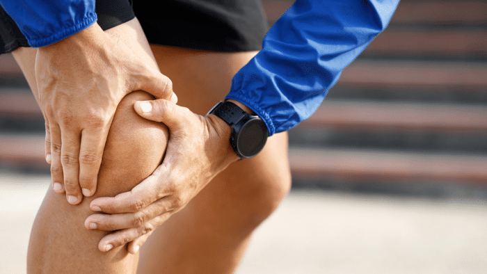Close-up of a person wearing a blue jacket, clutching their right knee with both hands, possibly experiencing pain or discomfort. They are outdoors, with blurred steps in the background.