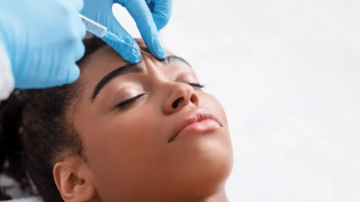 A person with closed eyes receives a cosmetic injection in the forehead from a medical professional wearing blue gloves. The focus is on the person's peaceful expression and the procedure being performed.