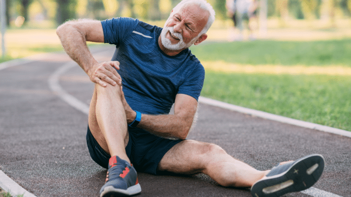 An older man sitting on a running track, holding his knee with a pained expression. He wears a blue t-shirt, black shorts, and red sneakers. The background shows a park with green grass and a blurred figure in the distance.