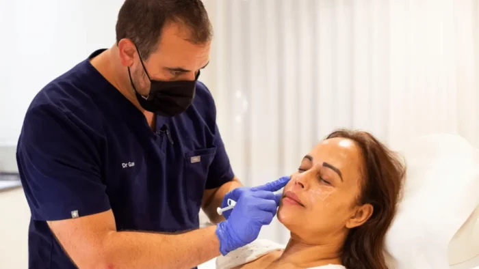 A person in a dark mask and gloves is administering a cosmetic procedure to a woman lying down. The woman appears relaxed, and the setting is a bright, clinical room.