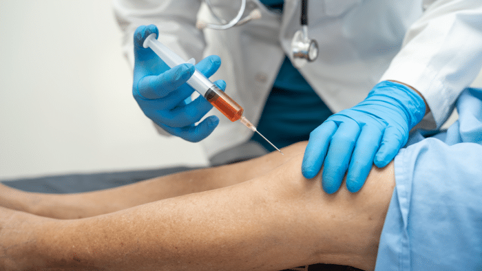 A medical professional in a white coat and blue gloves is administering an injection to a patient's knee. The syringe contains a reddish fluid, and the patient is lying down, covered partially by a blue cloth.
