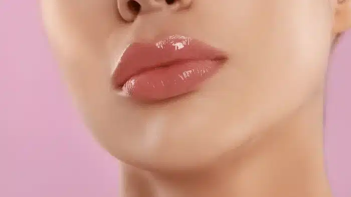 Close-up of a person’s glossy pink lips against a soft pink background, highlighting smooth skin and a slightly open mouth.