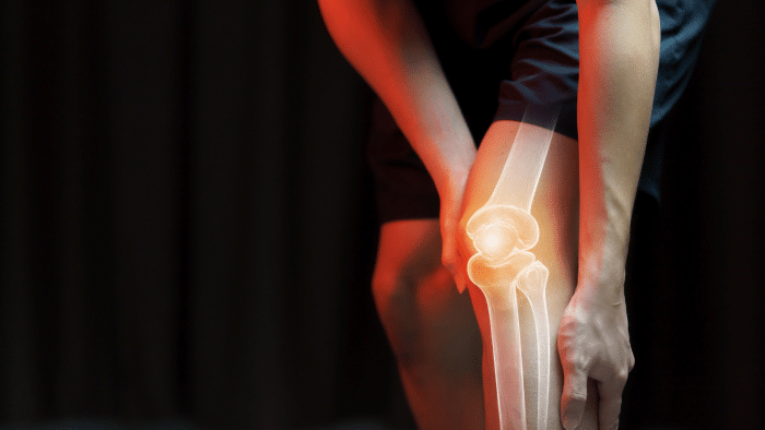 A person holding their knee, with a glowing overlay showing the knee joint and bones, suggesting joint pain or knee issues. The background is dark, highlighting the focus on the knee area.