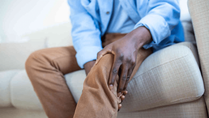 A person sitting on a couch, wearing brown pants and a blue shirt, is holding their knee with one hand, possibly indicating discomfort or pain. The focus is on the hand and knee, suggesting an issue with the leg.