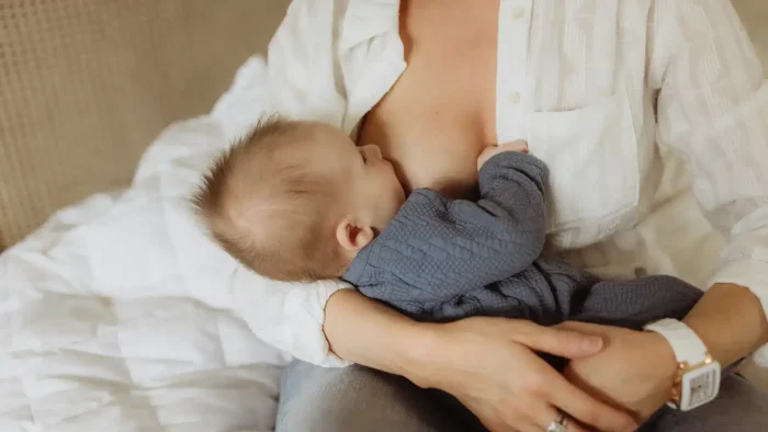 A baby wearing a blue outfit is being breastfed by a person wearing a white shirt. The scene is cozy, with a soft blanket in the background. The person's face is not visible, and they are wearing a watch on their wrist.