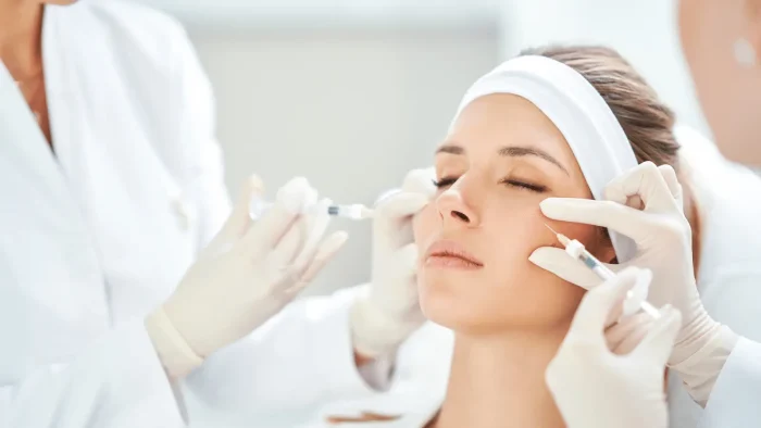 A person receives cosmetic facial injections from two practitioners. They are wearing a white headband, and the practitioners are dressed in white coats and gloves. The setting appears clinical or medical.