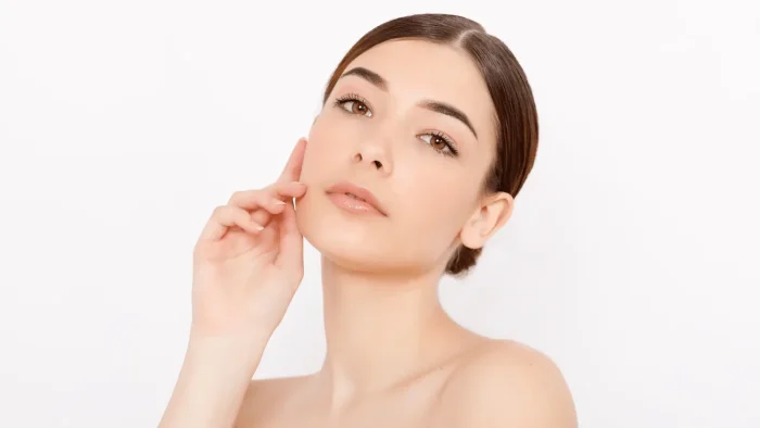 A woman with smooth skin and straight dark hair pulled back, gently touching her cheek with her fingers. She has a neutral expression and a plain white background.