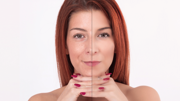 A close-up of a woman's face divided down the middle to show a makeup transformation. The left side of her face is natural with freckles and minimal makeup, while the right side is fully made up with even skin tone, enhanced eyes, and defined eyebrows. She has red hair and rests her chin on her hands.