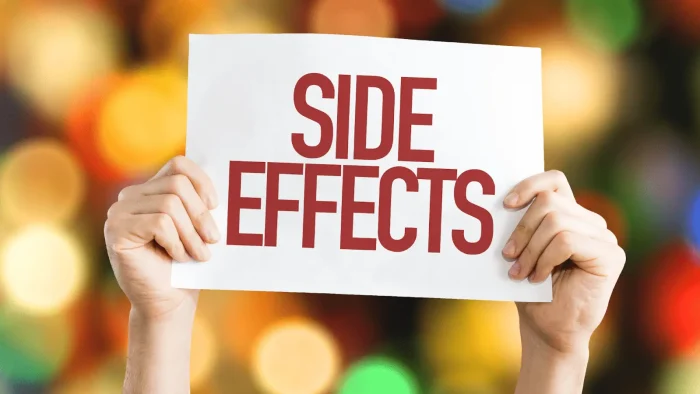 Two hands holding a white sign with the words "SIDE EFFECTS" in bold red letters. The background features blurred colorful lights, creating a festive atmosphere.