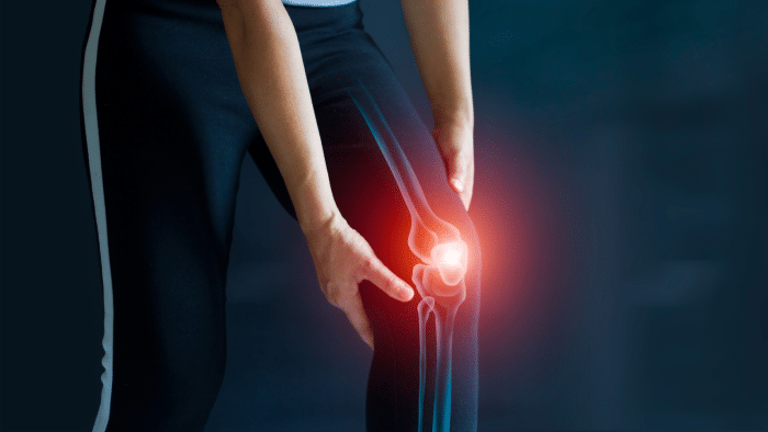A person holds their knee, with a glowing red area indicating pain or injury. The person is wearing dark athletic pants, and the background is blurred in a dark blue shade. An illustration of a knee joint is superimposed over the knee.