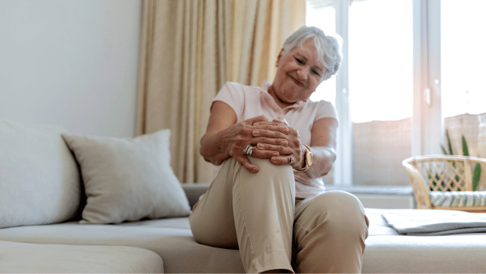 An elderly woman sitting on a couch indoors, holding her knee with both hands and showing a pained expression. She is wearing a light pink shirt and beige pants. A woven chair and large window with beige curtains are in the background.