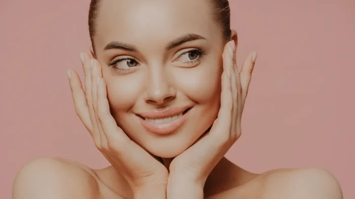 A woman with smooth skin smiles gently while holding her hands to her face. She is against a pink background, and her hair is pulled back, highlighting her natural beauty and serene expression.