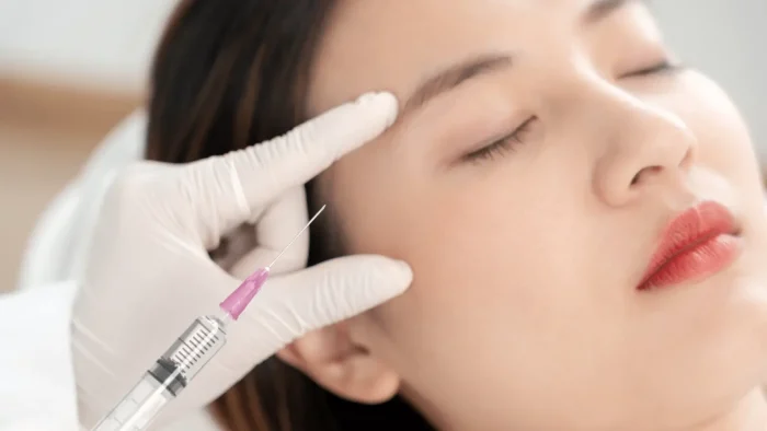 A person lying down with closed eyes is receiving an injection near the eyebrow. A gloved hand is seen holding the syringe. The setting appears to be a clinical or cosmetic procedure.
