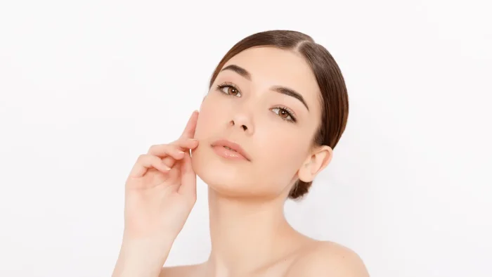 A woman with dark hair is gently touching her face. She has light makeup, natural-looking skin, and is gazing towards the camera against a plain white background.