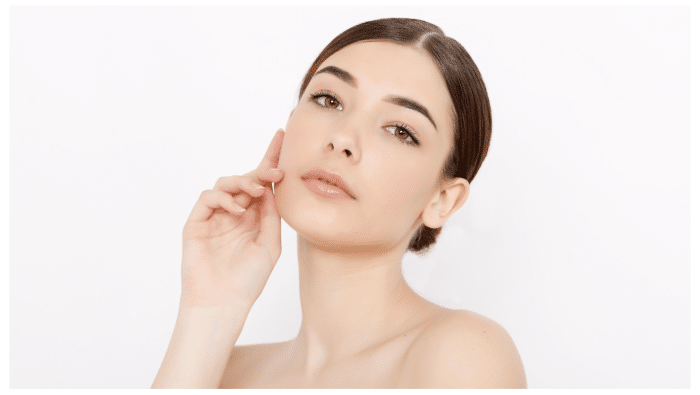 A person with fair skin and brown hair is posing against a plain white background. Their hair is neatly tied back, and they are wearing natural makeup. They are touching their cheek with one hand and looking directly at the camera with a neutral expression.