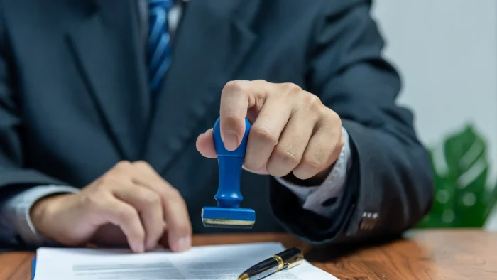 A person in a suit presses a blue stamp onto a document on a wooden desk. A pen lies beside the document. The scene suggests the approval or validation of paperwork in an office setting.