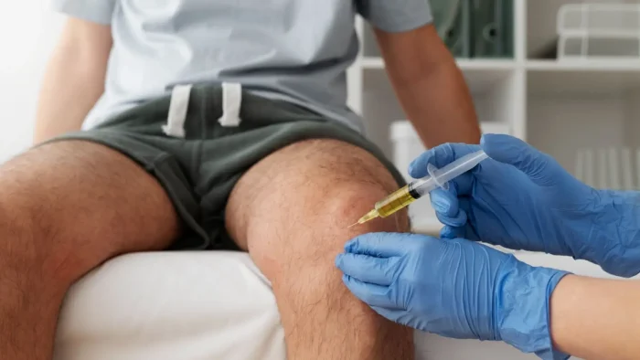 A person sitting on an examination table receives a knee injection from a healthcare worker wearing blue gloves. The syringe contains a yellow liquid. The focus is on the procedure, with the upper body of the patient not visible.