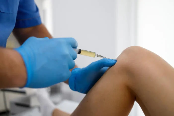 A healthcare professional wearing blue gloves is injecting a syringe into a patient's knee. The patient is seated, and the focus is on the knee and hands of the professional. The background is softly blurred.
