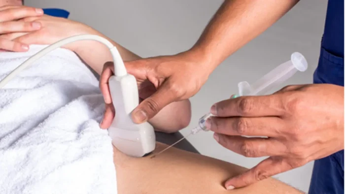 A medical professional performs an ultrasound-guided injection on a patient's abdomen. The patient is lying down, partially covered with a towel. The professional holds an ultrasound probe and a syringe.