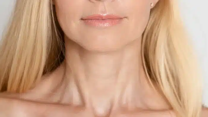 Close-up of a woman's lower face and neck, showcasing her lips and collarbone. She has long blonde hair, and her skin appears smooth and clear. The background is neutral, emphasizing her features.
