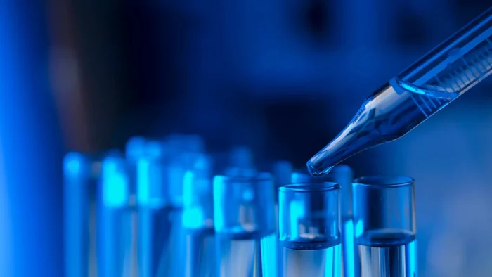 A close-up of a pipette dispensing liquid into a row of glass test tubes. The scene is bathed in a blue light, creating a scientific and laboratory atmosphere.