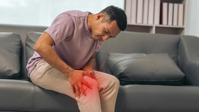 A man sitting on a couch holding his knee in pain, with a red glow indicating discomfort. He is wearing a purple shirt and beige pants, and the background features a gray sofa and bookshelves.