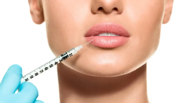 Close-up of a person with full lips, receiving a cosmetic lip injection. A gloved hand is holding a syringe near the lower lip. The background is neutral.