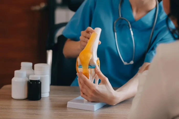 A healthcare professional in blue scrubs, wearing a stethoscope, explains a knee joint model to a patient. Various medication bottles are on the table in front of them.