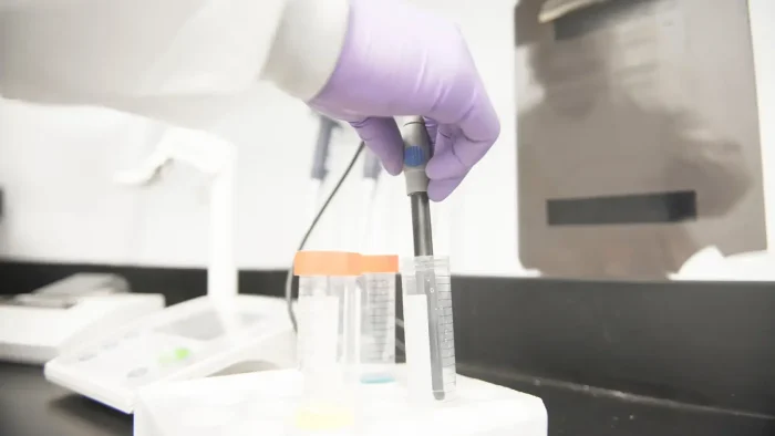 A gloved hand holding a capped test tube over a rack with various other test tubes in a lab setting. The background features lab equipment with a clipboard on the wall.