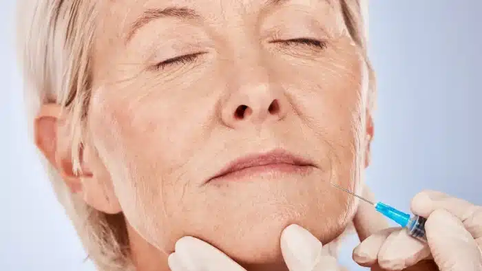 An older person with closed eyes receives a facial injection from a gloved hand, possibly a cosmetic procedure. The background is a soft blue, highlighting the subject's expression and the precision of the needle placement.