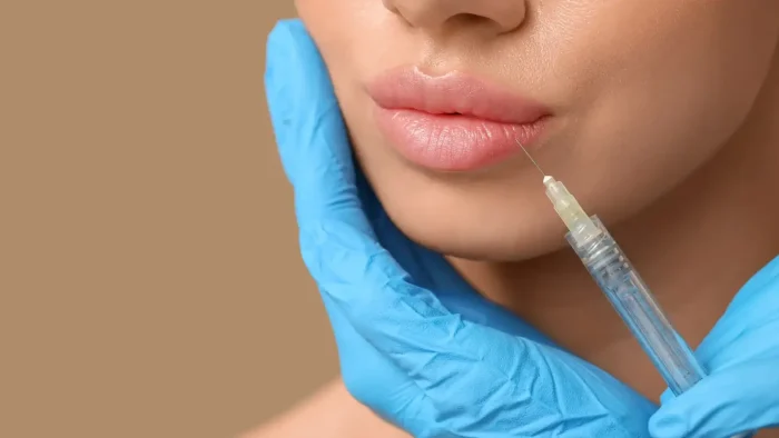 A person receiving a lip injection. A gloved hand gently holds the person's chin while a syringe approaches their lips. The background is a neutral beige color.